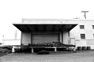 An abandoned loading dock attached to an old, worn-down building. The structure has a large metal shutter and a canopy overhead. The foreground is marked by cracked asphalt and scattered debris. In the background, there are utility poles and a glimpse of a stadium.