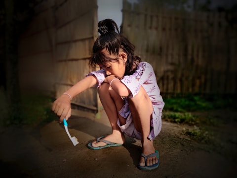 A young girl squats outdoors holding a toothbrush with toothpaste, wearing a long-sleeve dress and flip-flops. The background features a wooden fence and some greenery, with light and shadow creating a warm atmosphere.
