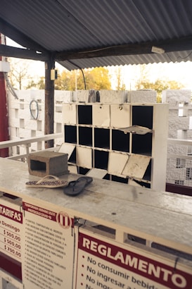 An empty kiosk or service counter with a partitioned shelf in the background, showing some wear. A concrete block and a pair of flip-flops are placed on the counter. Notices and regulations in Spanish are displayed in the foreground.