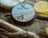 A tin of beard balm labeled 'Daily Grind Beard Co.' is placed on sandy ground, surrounded by driftwood and a halved lemon. The design on the tin features sunglasses on a sandy beach backdrop.