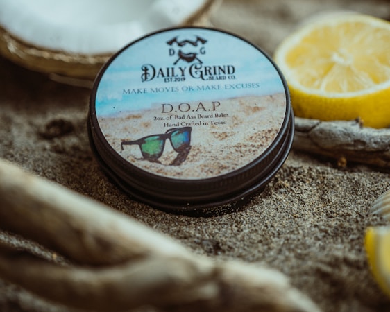 A tin of beard balm labeled 'Daily Grind Beard Co.' is placed on sandy ground, surrounded by driftwood and a halved lemon. The design on the tin features sunglasses on a sandy beach backdrop.