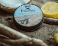 A tin of beard balm labeled 'Daily Grind Beard Co.' is placed on sandy ground, surrounded by driftwood and a halved lemon. The design on the tin features sunglasses on a sandy beach backdrop.
