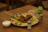 A large metal platter filled with rice and meat garnished with slices of onion, green bell pepper, cucumber, and carrot sits on a wooden table. Accompanying the meal is a bottle of Mountain Dew, a small bowl of sauce, and a bowl containing a white liquid, likely yogurt or raita. Salt and pepper shakers are also present on the table.