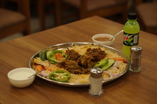 A large metal platter filled with rice and meat garnished with slices of onion, green bell pepper, cucumber, and carrot sits on a wooden table. Accompanying the meal is a bottle of Mountain Dew, a small bowl of sauce, and a bowl containing a white liquid, likely yogurt or raita. Salt and pepper shakers are also present on the table.