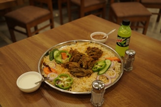 A large stainless steel platter filled with a rice dish garnished with slices of green bell peppers, onion rings, and tomatoes is placed on a wooden table. In the center of the dish, there are pieces of spiced meat. Accompanying the meal are a small bowl of white sauce with a spoon, a smaller bowl of reddish condiment with a spoon, a bottle of Mountain Dew, and two salt and pepper shakers. In the background, there are wooden chairs around the table.