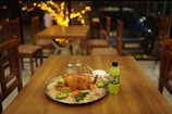 A table in a cozy restaurant setting features a large tray with a roasted chicken on a bed of rice, garnished with sliced green peppers, carrots, and onions. Beside the platter, there are salt and pepper shakers, a bowl with white sauce, and a bottle of Mountain Dew. In the background, wooden chairs and tables are visible, along with ambient warm lighting from string lights.