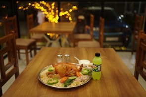A table in a cozy restaurant setting features a large tray with a roasted chicken on a bed of rice, garnished with sliced green peppers, carrots, and onions. Beside the platter, there are salt and pepper shakers, a bowl with white sauce, and a bottle of Mountain Dew. In the background, wooden chairs and tables are visible, along with ambient warm lighting from string lights.