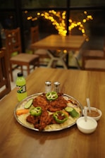 A wooden table is set with a large platter of grilled chicken pieces garnished with slices of green bell peppers, cucumbers, and onions, over a bed of rice. Accompanying the dish are small bowls of sauces and a bottle of green soda. In the background, soft, warm lighting creates a cozy dining ambiance.