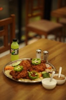 A platter filled with grilled meat pieces garnished with green bell peppers and served on a bed of rice with sliced cucumbers, carrots, and onions. On the side, there are two bowls containing sauces or dips, a bottle of Mountain Dew, and salt and pepper shakers. The setting appears to be a restaurant with wooden tables and chairs.