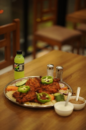 A platter filled with grilled meat pieces garnished with green bell peppers and served on a bed of rice with sliced cucumbers, carrots, and onions. On the side, there are two bowls containing sauces or dips, a bottle of Mountain Dew, and salt and pepper shakers. The setting appears to be a restaurant with wooden tables and chairs.