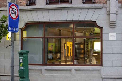 A Nikon store with a large window display features photographic equipment and art posters. A nearby green postal box is labeled 'China Post'. A blue sign indicates a traffic restriction, showing a car with a red line through it. The building facade is made of grey stone with a classical design.