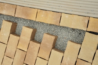 A pattern of rectangular beige bricks arranged on a layer of small gray gravel, interspersed with larger gaps. The surface appears to be under construction or preparation for a walkway or patio. The top edge features a lighter, lined surface, possibly for transitioning.