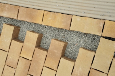 A pattern of rectangular beige bricks arranged on a layer of small gray gravel, interspersed with larger gaps. The surface appears to be under construction or preparation for a walkway or patio. The top edge features a lighter, lined surface, possibly for transitioning.