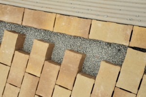 A pattern of rectangular beige bricks arranged on a layer of small gray gravel, interspersed with larger gaps. The surface appears to be under construction or preparation for a walkway or patio. The top edge features a lighter, lined surface, possibly for transitioning.