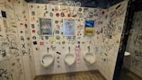 The tiled walls of a restroom are covered in a variety of graffiti, stickers, and drawings, creating a chaotic and urban look. Three urinals are lined up along the wall, situated below the graffiti. There are several different colors and styles of writing, contributing to the cluttered appearance.
