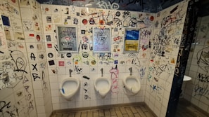 The tiled walls of a restroom are covered in a variety of graffiti, stickers, and drawings, creating a chaotic and urban look. Three urinals are lined up along the wall, situated below the graffiti. There are several different colors and styles of writing, contributing to the cluttered appearance.