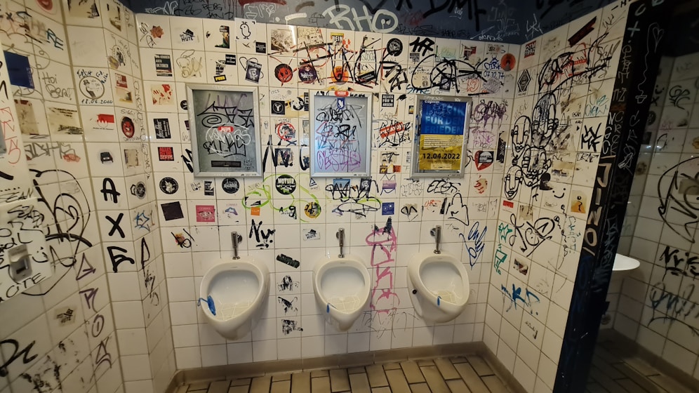 The tiled walls of a restroom are covered in a variety of graffiti, stickers, and drawings, creating a chaotic and urban look. Three urinals are lined up along the wall, situated below the graffiti. There are several different colors and styles of writing, contributing to the cluttered appearance.