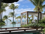 A serene tropical setting featuring lush green palm trees surrounding a luxurious pool. In the background, the blue ocean meets the sky. Wooden cabanas with white curtains are positioned near the water, offering a relaxing environment.