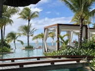 A serene tropical setting featuring lush green palm trees surrounding a luxurious pool. In the background, the blue ocean meets the sky. Wooden cabanas with white curtains are positioned near the water, offering a relaxing environment.