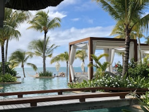A serene tropical setting featuring lush green palm trees surrounding a luxurious pool. In the background, the blue ocean meets the sky. Wooden cabanas with white curtains are positioned near the water, offering a relaxing environment.