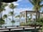 A serene tropical setting featuring lush green palm trees surrounding a luxurious pool. In the background, the blue ocean meets the sky. Wooden cabanas with white curtains are positioned near the water, offering a relaxing environment.