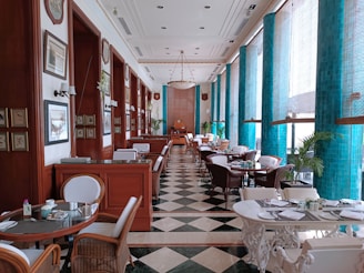 An elegant dining area adorned with floor-to-ceiling turquoise tiles and large windows allowing ample natural light. The space features a checkered black and white floor, wooden paneling with framed paintings, and a series of round tables with wicker chairs. There are also plush booths and green potted plants adding to the luxurious ambiance.