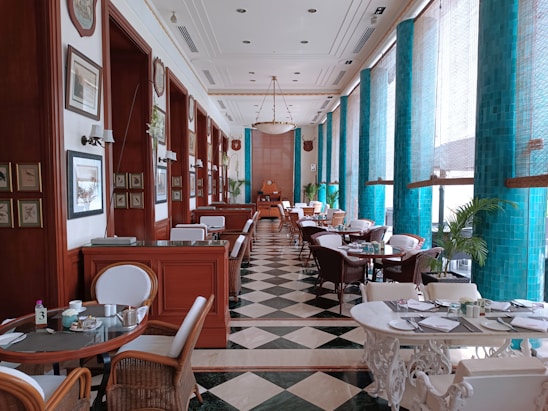 An elegant dining area adorned with floor-to-ceiling turquoise tiles and large windows allowing ample natural light. The space features a checkered black and white floor, wooden paneling with framed paintings, and a series of round tables with wicker chairs. There are also plush booths and green potted plants adding to the luxurious ambiance.