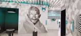 A stylized geometric mural of a man with glasses takes up most of a wall. The background features a geometric pattern in shades of gray and white, alongside turquoise accents. Above, there are floating installations resembling stylized birds. The text 'Reality is merely' is prominently displayed in turquoise against a white wall. Additional decorative elements and interactive exhibits are found around the room.