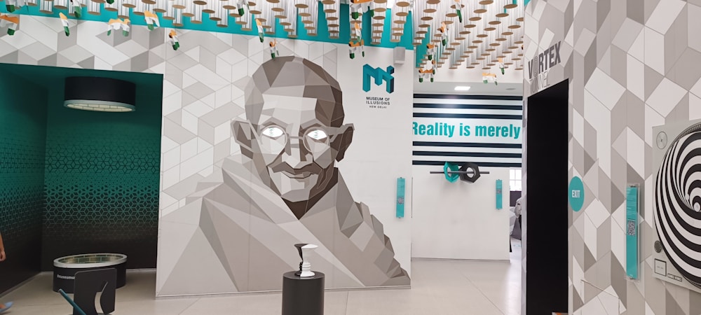 A stylized geometric mural of a man with glasses takes up most of a wall. The background features a geometric pattern in shades of gray and white, alongside turquoise accents. Above, there are floating installations resembling stylized birds. The text 'Reality is merely' is prominently displayed in turquoise against a white wall. Additional decorative elements and interactive exhibits are found around the room.
