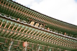 An intricately decorated traditional Korean roof with vibrant green, red, and white patterns. The detailed artwork includes floral motifs and geometric designs. A wooden sign with Korean script is prominently displayed.