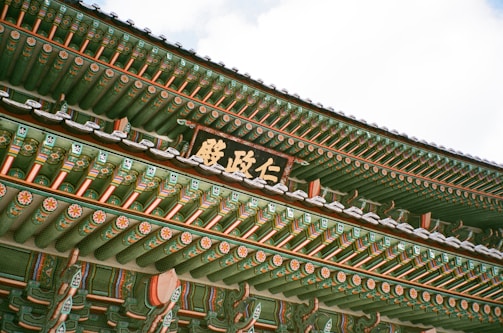 An intricately decorated traditional Korean roof with vibrant green, red, and white patterns. The detailed artwork includes floral motifs and geometric designs. A wooden sign with Korean script is prominently displayed.