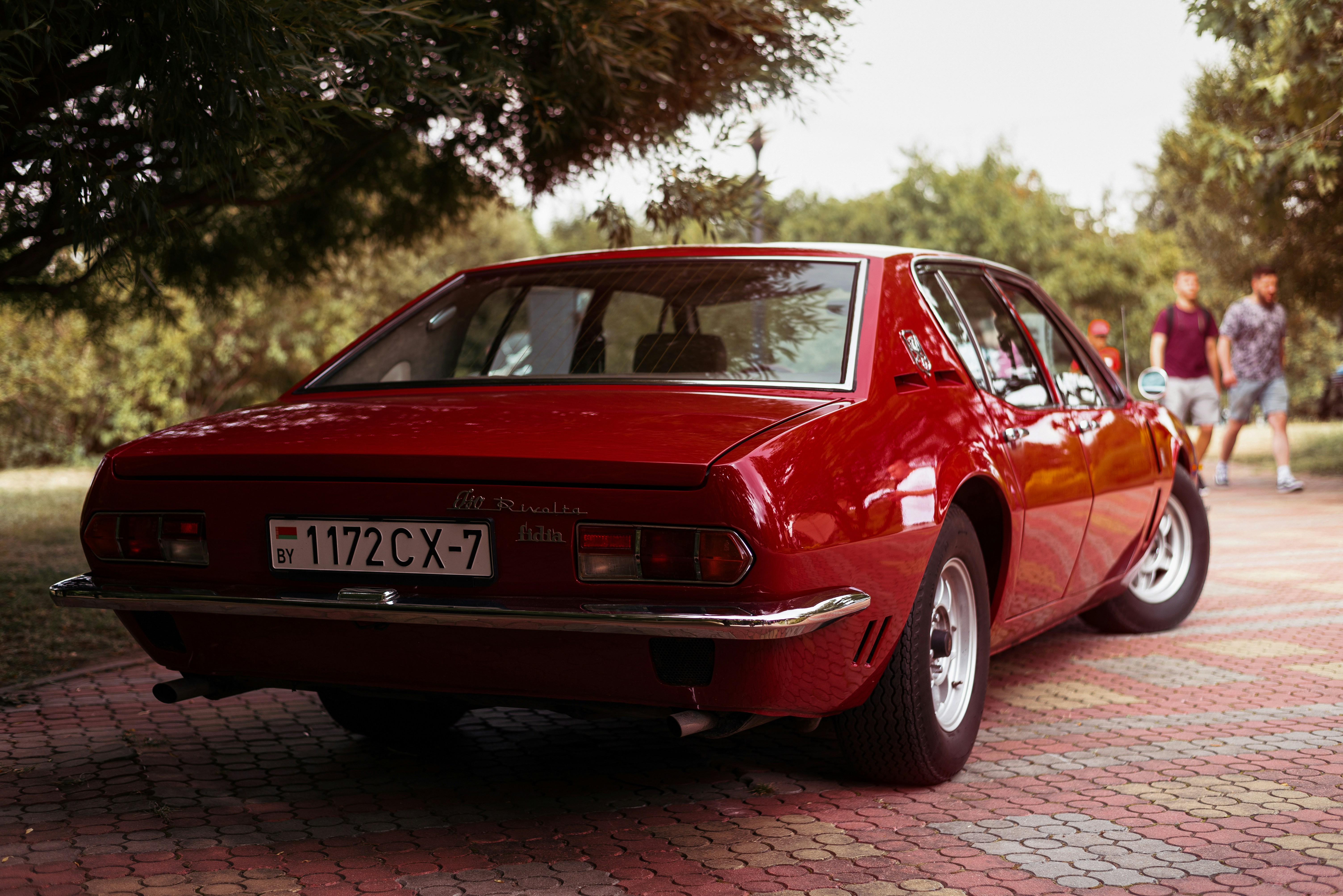 a red car parked on a brick road with people walking by