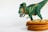 A green toy dinosaur with orange and yellow accents is positioned above a stack of freshly baked cookies with visible chocolate chips. The dinosaur appears to be looking at the cookies against a plain white background.
