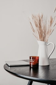 A minimalistic setup on a dark wooden table featuring a closed laptop, a brown candle labeled 'Orange & Blossom,' and a white ceramic pitcher filled with dry wheat stalks against a plain light-colored wall.