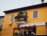 A two-story building with a warm yellow facade, featuring wooden shutters on the windows. There is a small balcony adorned with plants and flowers, and an Italian flag is hanging from the wall near the window.