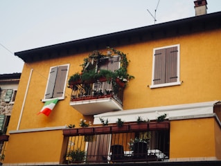 A two-story building with a warm yellow facade, featuring wooden shutters on the windows. There is a small balcony adorned with plants and flowers, and an Italian flag is hanging from the wall near the window.