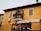 A two-story building with a warm yellow facade, featuring wooden shutters on the windows. There is a small balcony adorned with plants and flowers, and an Italian flag is hanging from the wall near the window.