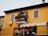 A two-story building with a warm yellow facade, featuring wooden shutters on the windows. There is a small balcony adorned with plants and flowers, and an Italian flag is hanging from the wall near the window.