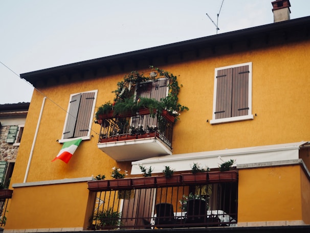 A two-story building with a warm yellow facade, featuring wooden shutters on the windows. There is a small balcony adorned with plants and flowers, and an Italian flag is hanging from the wall near the window.