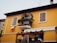 A two-story building with a warm yellow facade, featuring wooden shutters on the windows. There is a small balcony adorned with plants and flowers, and an Italian flag is hanging from the wall near the window.