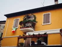 A two-story building with a warm yellow facade, featuring wooden shutters on the windows. There is a small balcony adorned with plants and flowers, and an Italian flag is hanging from the wall near the window.