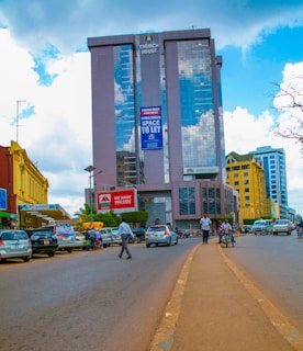 A bustling urban street scene with a tall glass skyscraper named 'Church House' reflecting clouds in its facade. Various people and vehicles fill the street, and there's a prominent sign reading 'We Want You Safe'. Buildings of different colors line the street, enhancing the vibrant urban landscape.
