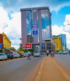 A bustling urban street scene with a tall glass skyscraper named 'Church House' reflecting clouds in its facade. Various people and vehicles fill the street, and there's a prominent sign reading 'We Want You Safe'. Buildings of different colors line the street, enhancing the vibrant urban landscape.