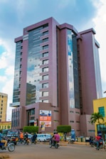 A tall modern building with reflective glass windows and a sign that reads 'CHURH HOUS' stands prominently in an urban area. The ground level has visible advertisements and multiple signs for businesses, including one for 'Equity Bank'. In front of the building, there is a bustling street with motorcyclists, pedestrians, and vehicles. Managed bushes and a small palm tree add greenery to the scene, while clouds dot a bright blue sky.