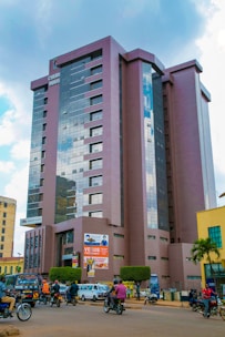 A tall modern building with reflective glass windows and a sign that reads 'CHURH HOUS' stands prominently in an urban area. The ground level has visible advertisements and multiple signs for businesses, including one for 'Equity Bank'. In front of the building, there is a bustling street with motorcyclists, pedestrians, and vehicles. Managed bushes and a small palm tree add greenery to the scene, while clouds dot a bright blue sky.