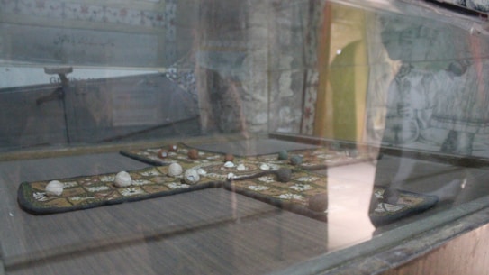 A glass display case houses an ancient board game, featuring a cross-shaped wooden board with carved patterns. Small stones or game pieces are placed on the squares of the board. The background is a wall with faded murals or intricate designs, giving an antique and historical ambiance.