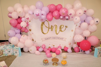 A festive arrangement features pink, purple, and white balloons surrounding a backdrop with 'One Happy Birthday' written in gold glitter letters. Decorations include small butterfly stickers on some balloons and tables laid out with gift bags and jars filled with colorful candies.