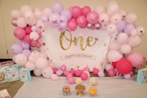 A festive arrangement features pink, purple, and white balloons surrounding a backdrop with 'One Happy Birthday' written in gold glitter letters. Decorations include small butterfly stickers on some balloons and tables laid out with gift bags and jars filled with colorful candies.