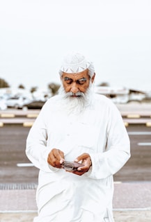 An elderly man with a white beard and traditional white attire is holding an open wallet. He is wearing a white cap and standing outdoors on a pavement with a road and vehicles blurred in the background.