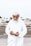 An elderly man with a white beard and traditional white attire is holding an open wallet. He is wearing a white cap and standing outdoors on a pavement with a road and vehicles blurred in the background.